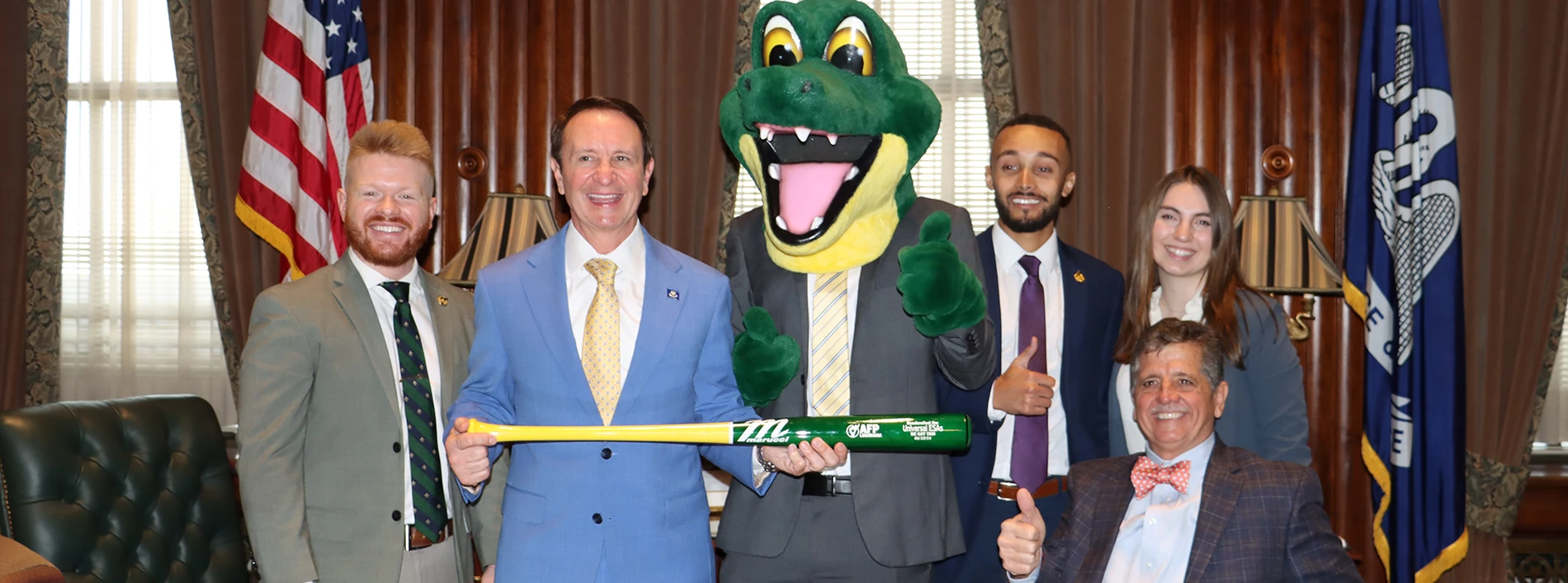 Governor Landry in a blue suit holds a green and yellow baseball bat. Four people and a green alligator mascot pose in a government office with U.S. and Louisiana flags.
